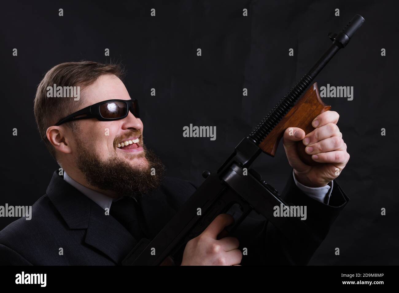 Homme à barbe mature en lunettes de soleil vêtu en costume avec tommy canon sur fond noir Banque D'Images