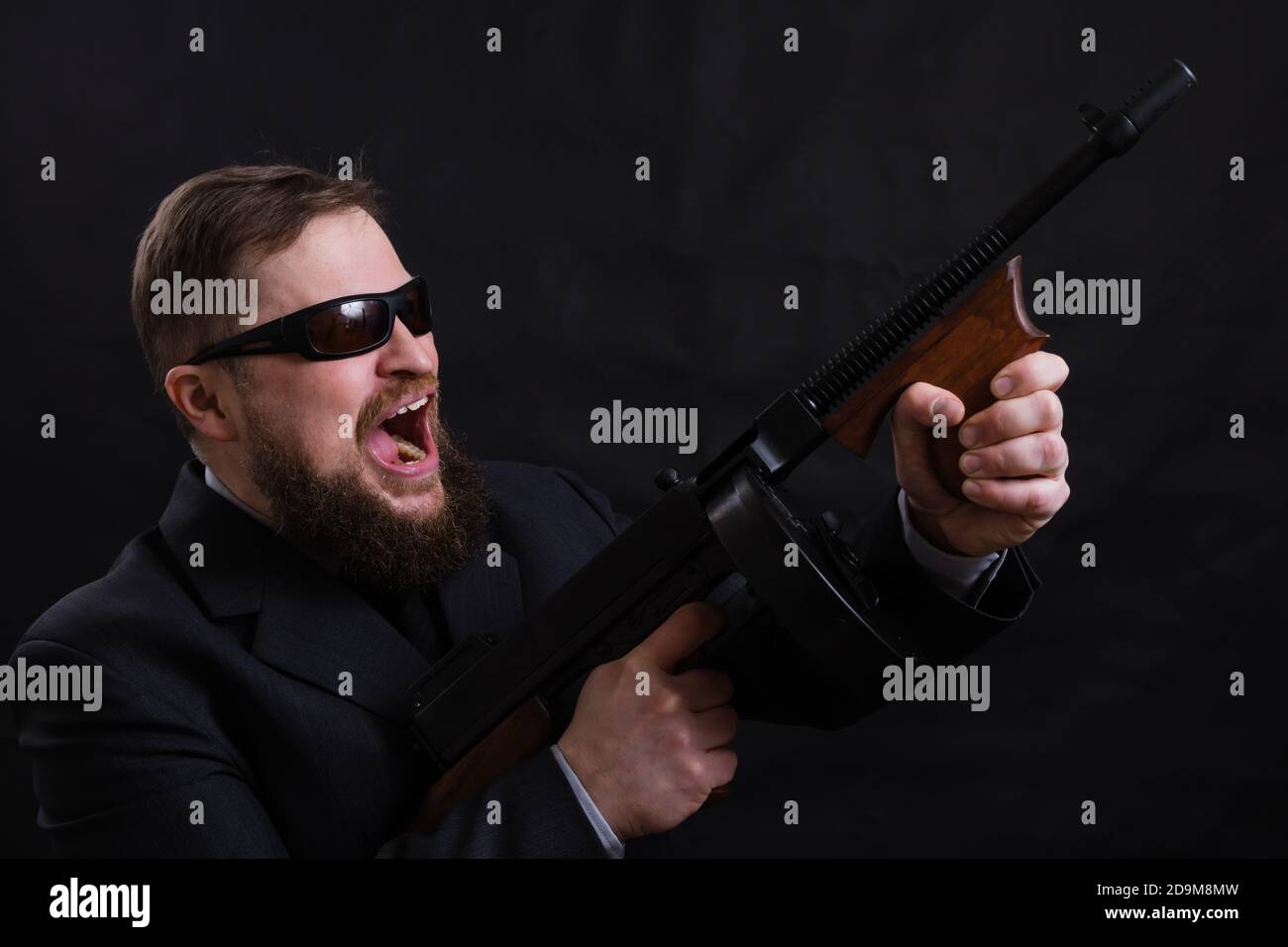 Homme à barbe mature en lunettes de soleil vêtu en costume avec tommy canon sur fond noir Banque D'Images