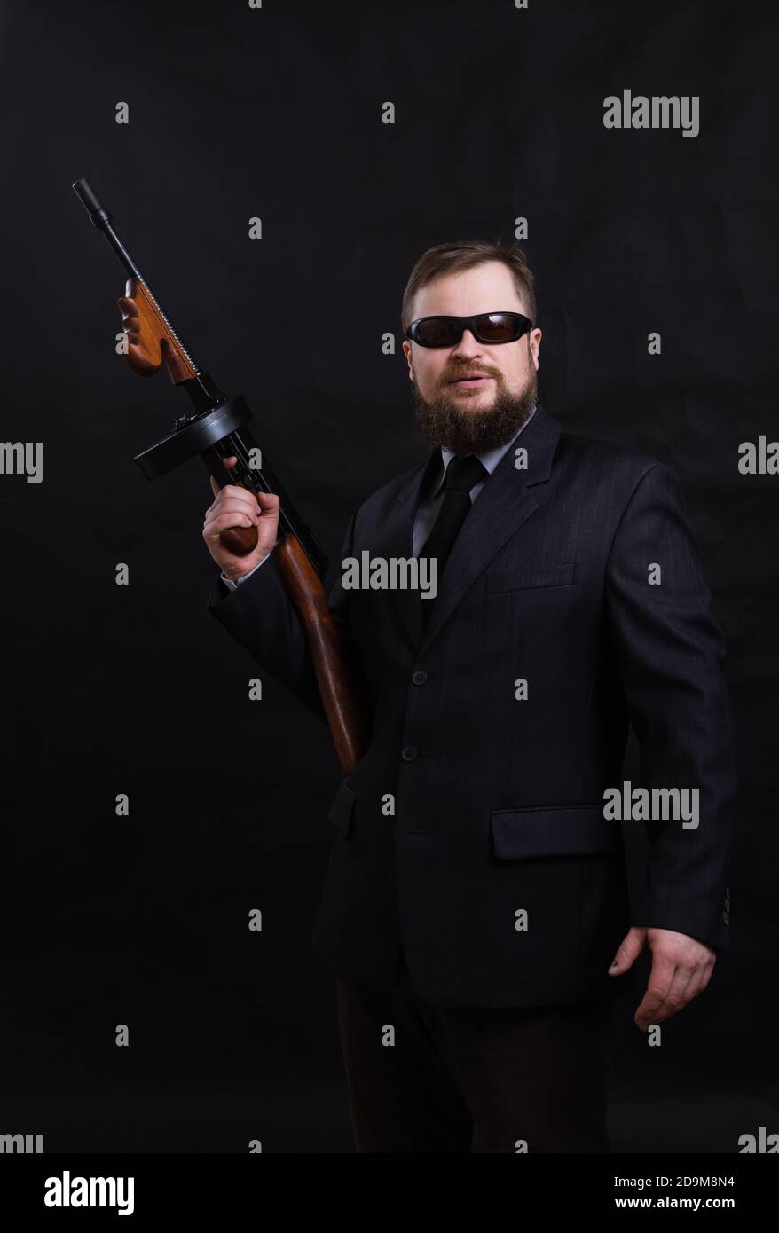 Homme à barbe mature en lunettes de soleil vêtu en costume avec tommy canon sur fond noir Banque D'Images
