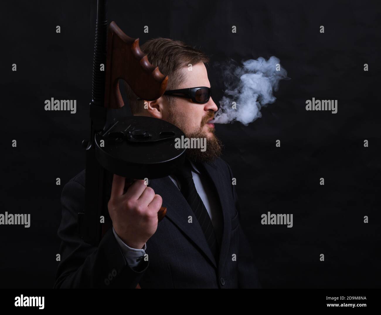 Homme à barbe mature en lunettes de soleil vêtu en costume avec tommy canon sur fond noir Banque D'Images