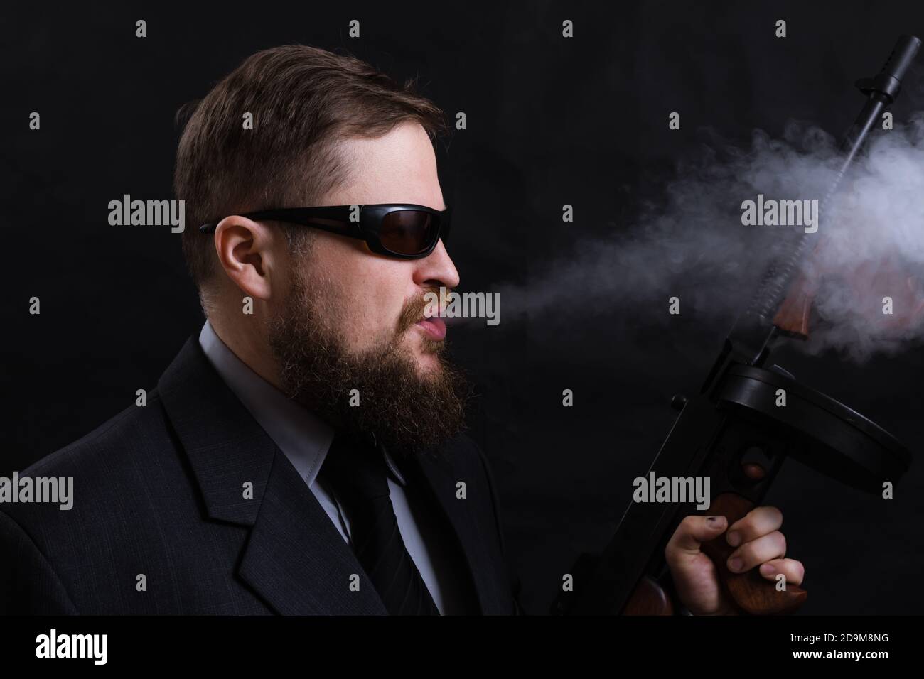 Homme à barbe mature en lunettes de soleil vêtu en costume avec tommy canon sur fond noir Banque D'Images