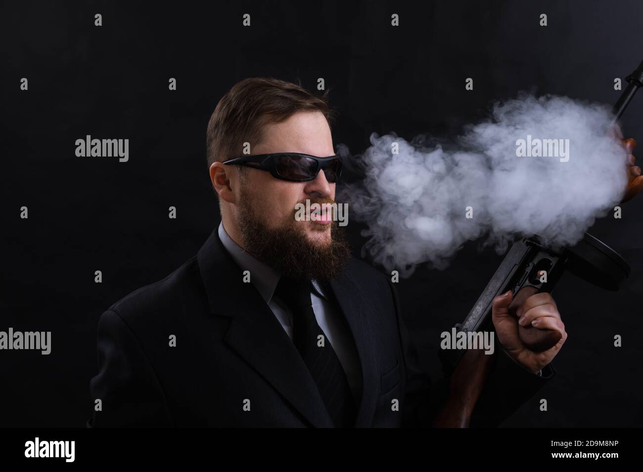 Homme à barbe mature en lunettes de soleil vêtu en costume avec tommy canon sur fond noir Banque D'Images