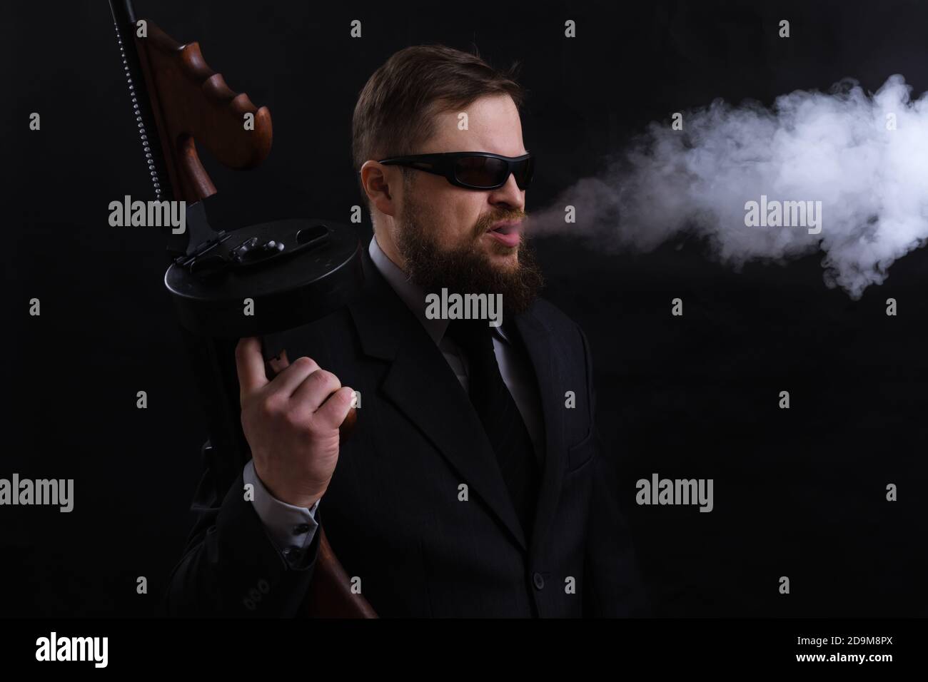 Homme à barbe mature en lunettes de soleil vêtu en costume avec tommy canon sur fond noir Banque D'Images