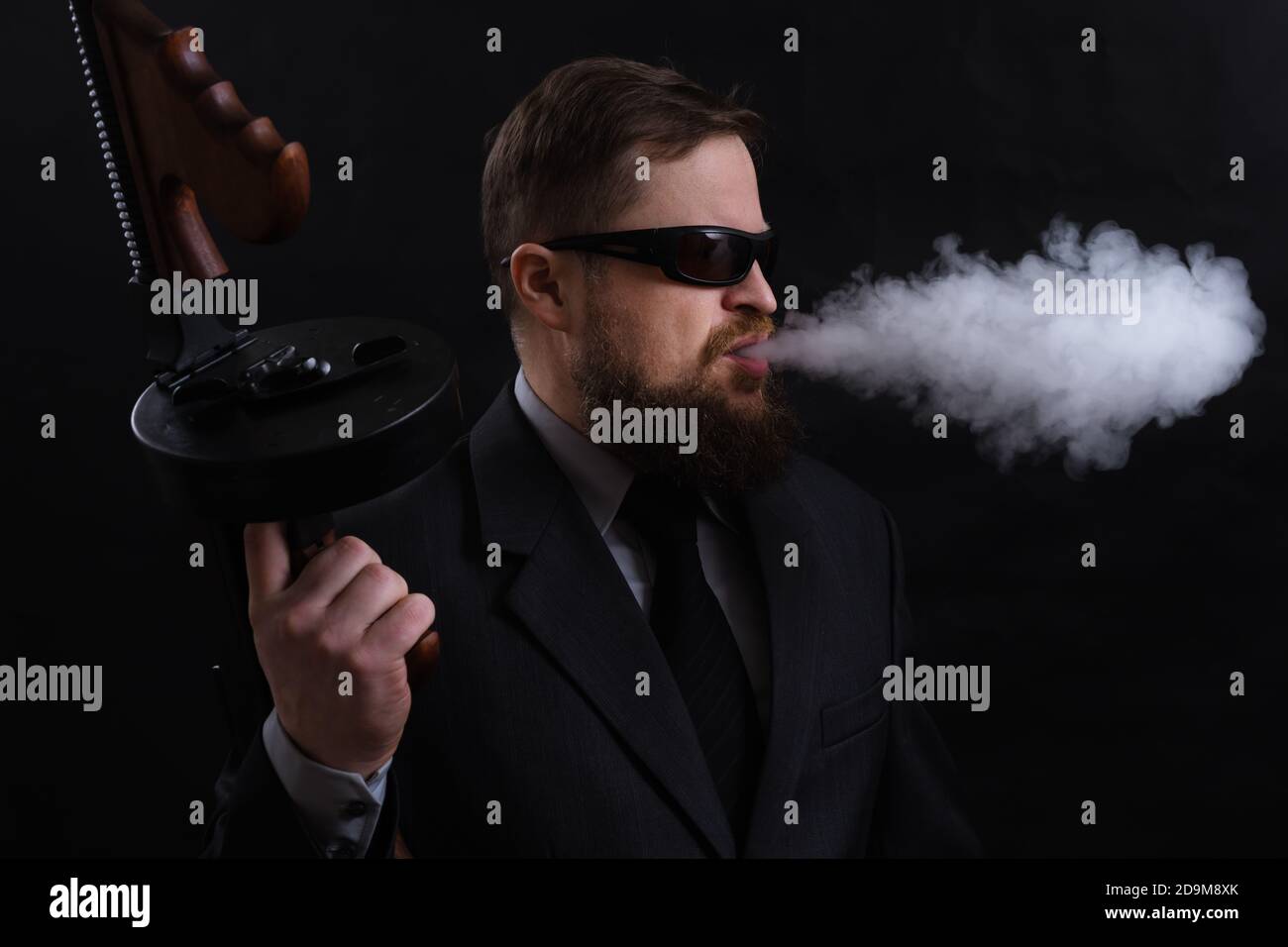 Homme à barbe mature en lunettes de soleil vêtu en costume avec tommy canon sur fond noir Banque D'Images