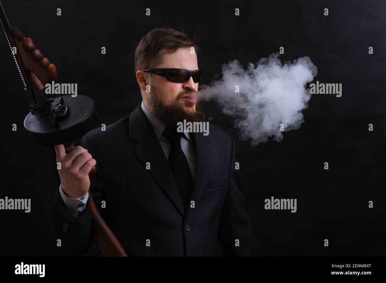 Homme à barbe mature en lunettes de soleil vêtu en costume avec tommy canon sur fond noir Banque D'Images