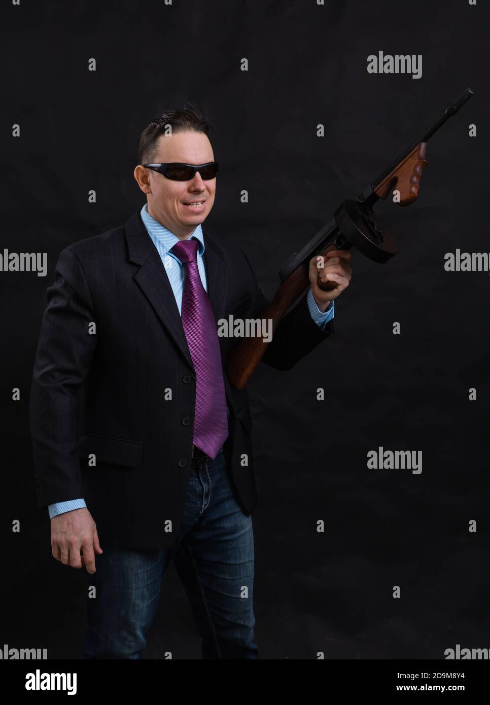 Gangster mature en lunettes de soleil vêtu en costume avec un fusil tommy sur fond noir Banque D'Images