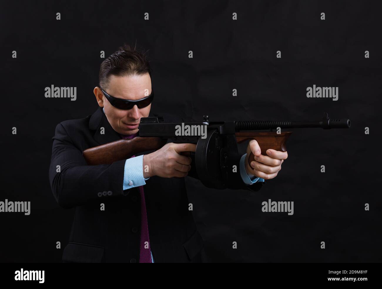 Gangster mature en lunettes de soleil vêtu en costume avec un fusil tommy sur fond noir Banque D'Images