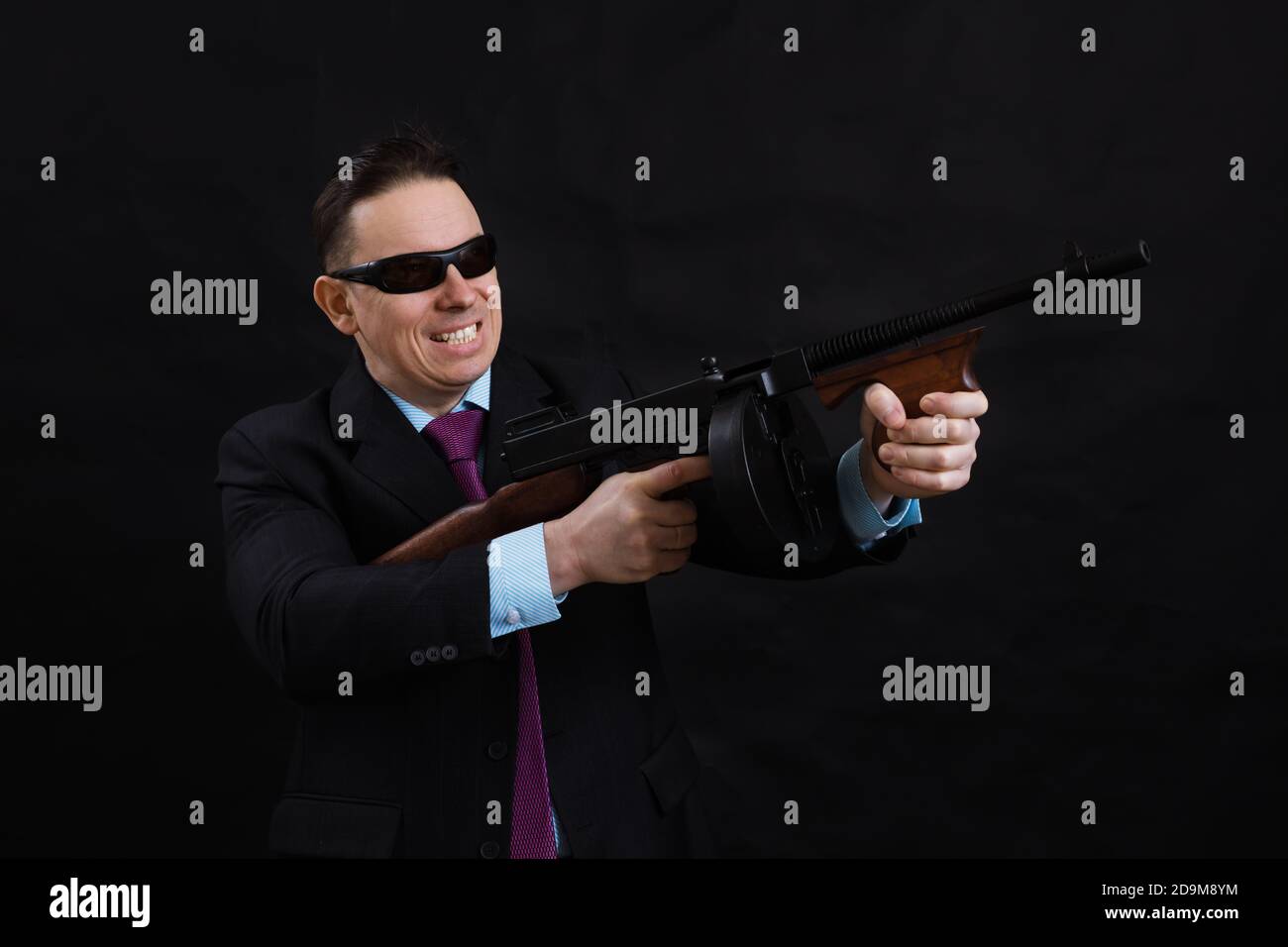 Gangster mature en lunettes de soleil vêtu en costume avec un fusil tommy sur fond noir Banque D'Images