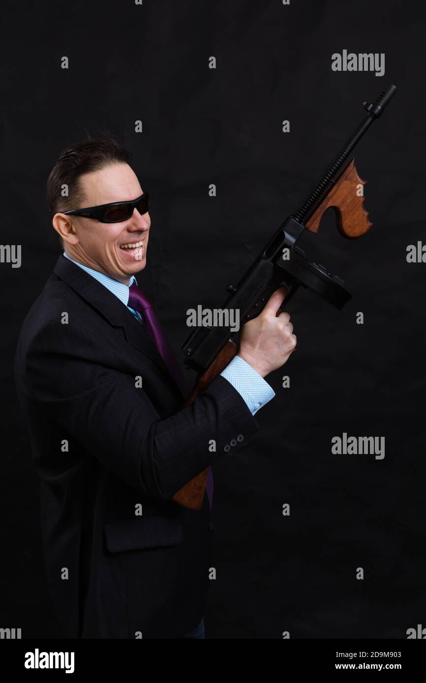 Gangster mature en lunettes de soleil vêtu en costume avec un fusil tommy sur fond noir Banque D'Images