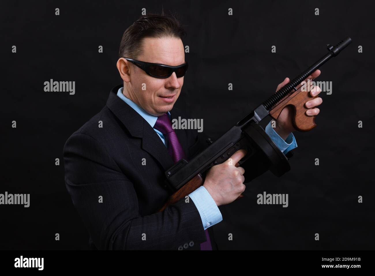 Gangster mature en lunettes de soleil vêtu en costume avec un fusil tommy sur fond noir Banque D'Images