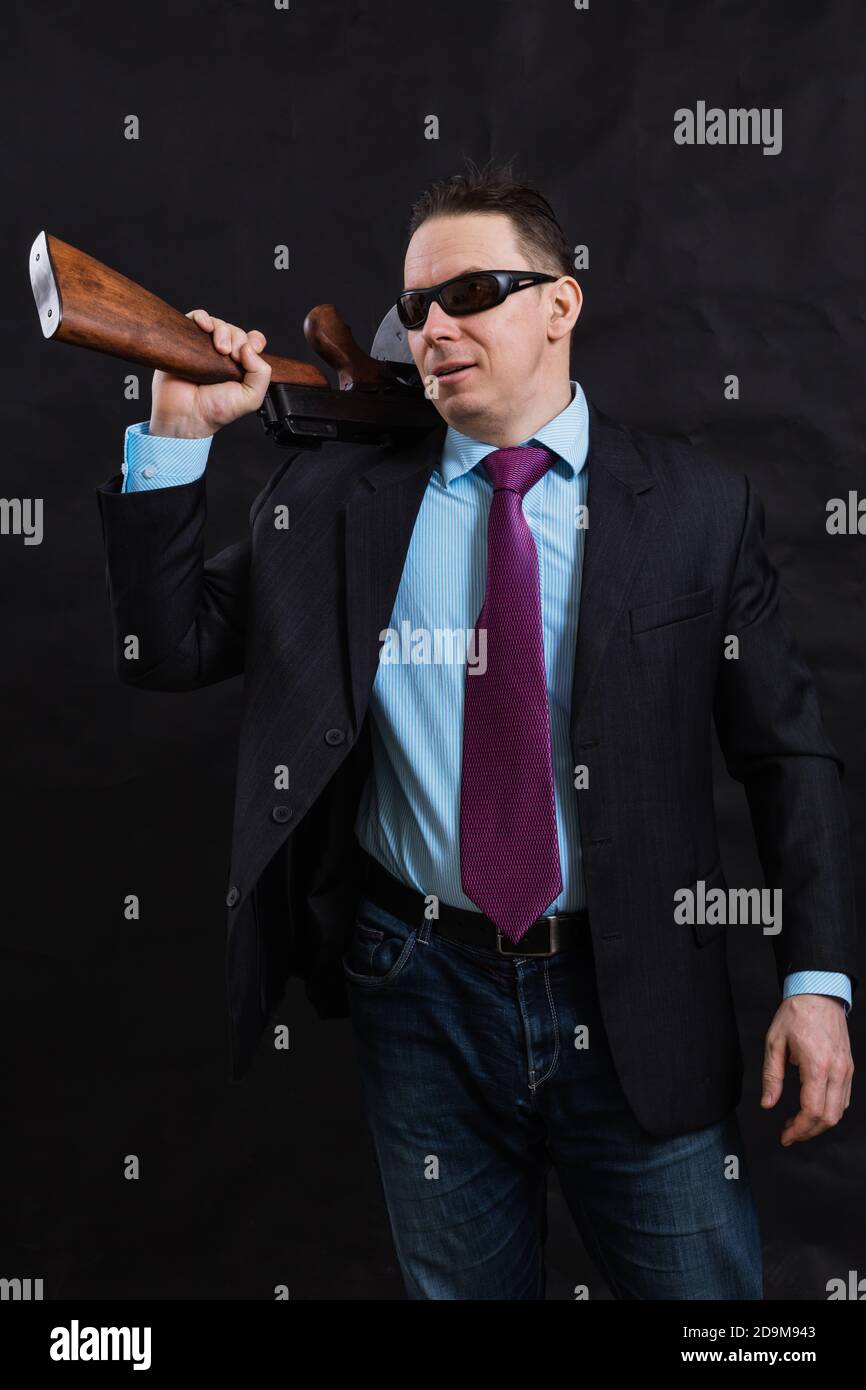 Gangster mature en lunettes de soleil vêtu en costume avec un fusil tommy sur fond noir Banque D'Images