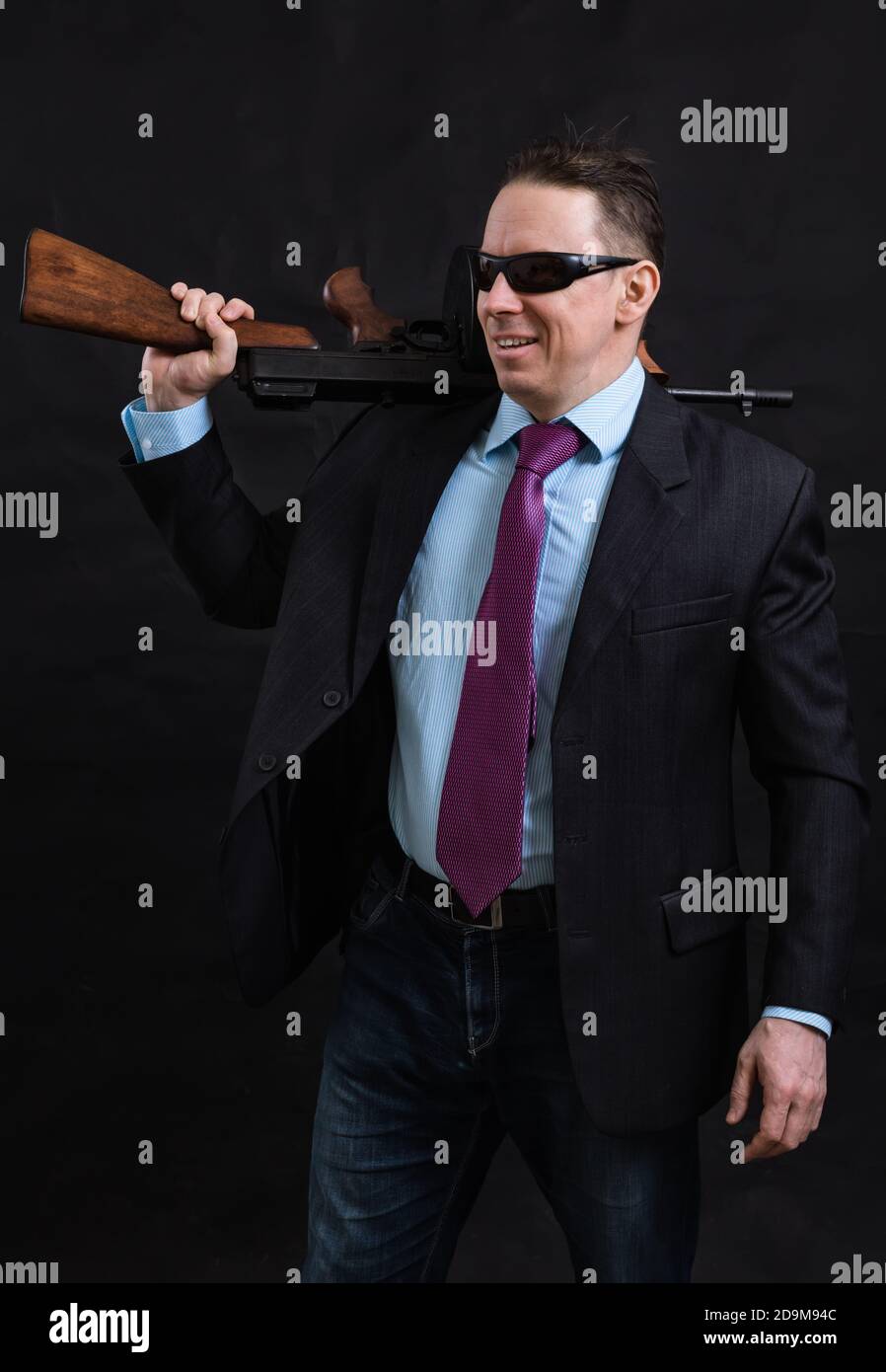 Gangster mature en lunettes de soleil vêtu en costume avec un fusil tommy sur fond noir Banque D'Images