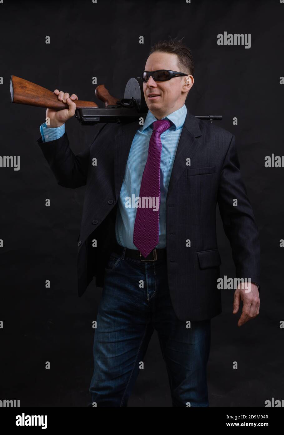 Gangster mature en lunettes de soleil vêtu en costume avec un fusil tommy sur fond noir Banque D'Images