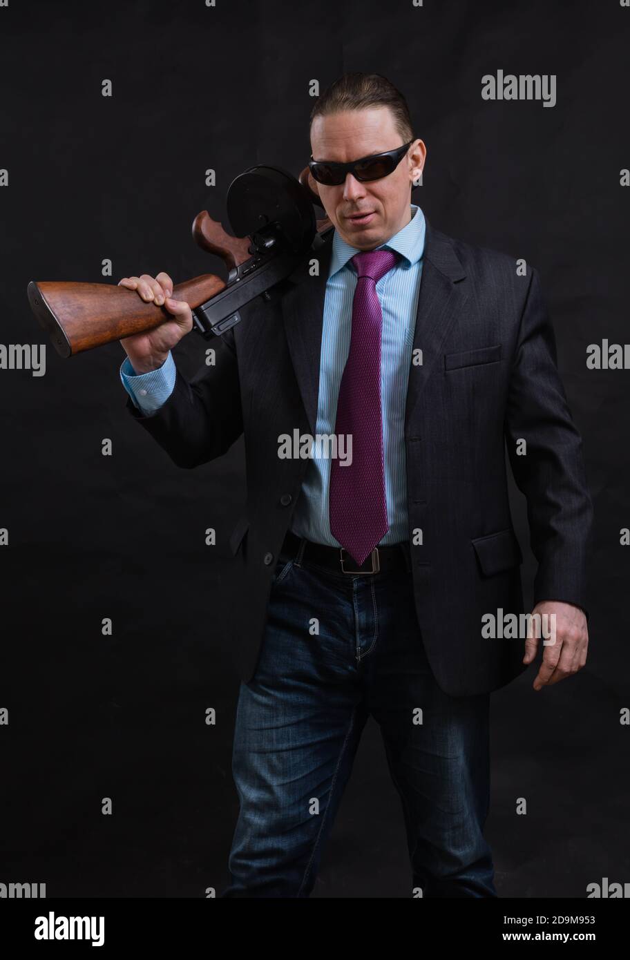 Gangster mature en lunettes de soleil vêtu en costume avec un fusil tommy sur fond noir Banque D'Images