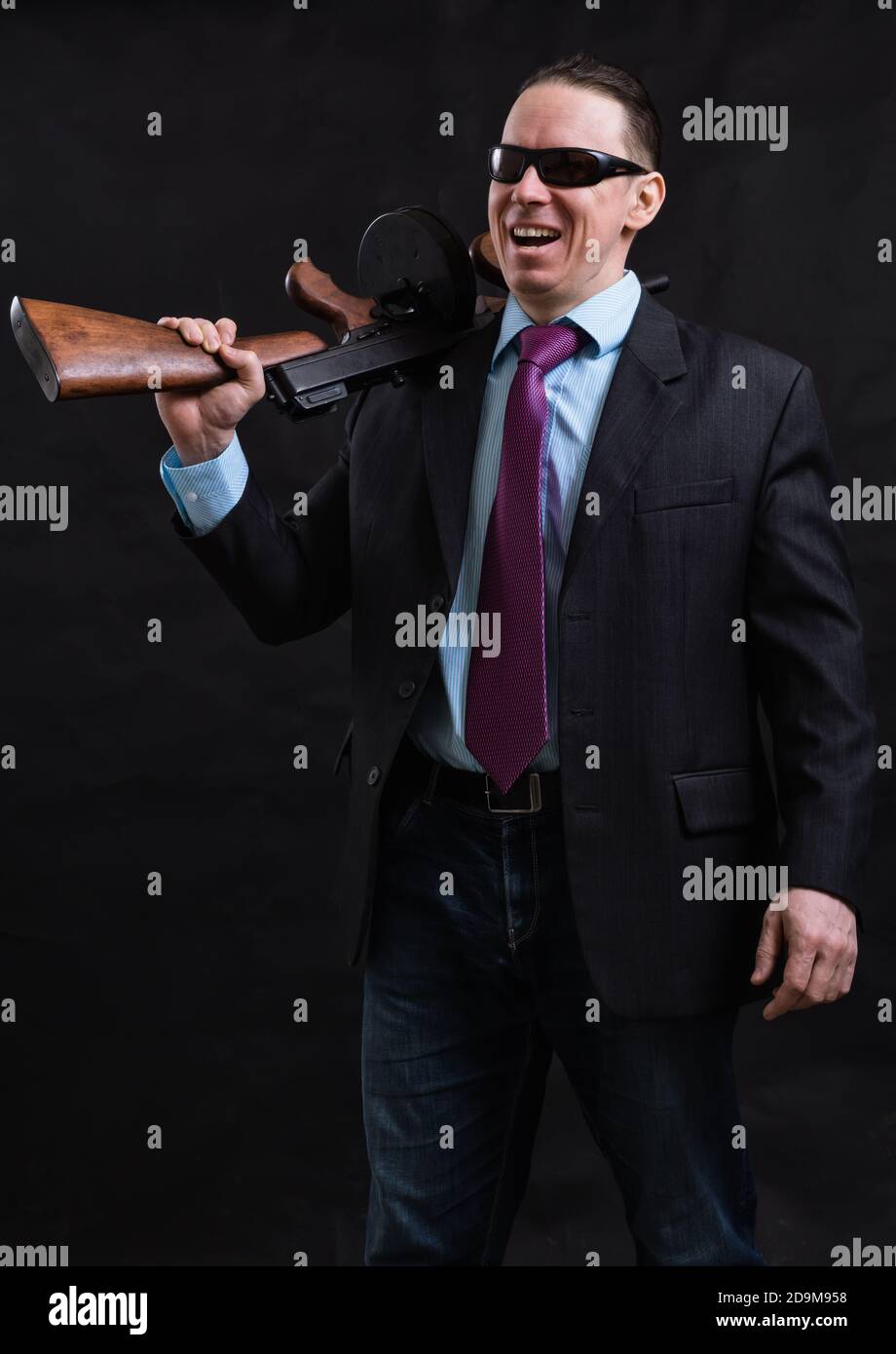 Gangster mature en lunettes de soleil vêtu en costume avec un fusil tommy sur fond noir Banque D'Images
