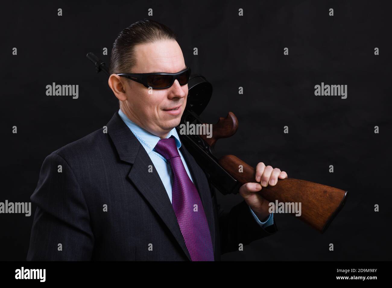 Gangster mature en lunettes de soleil vêtu en costume avec un fusil tommy sur fond noir Banque D'Images