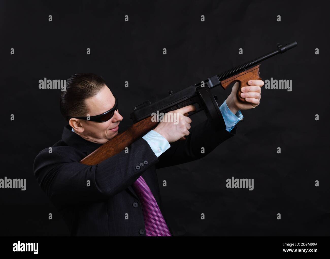 Gangster mature en lunettes de soleil vêtu en costume avec un fusil tommy sur fond noir Banque D'Images