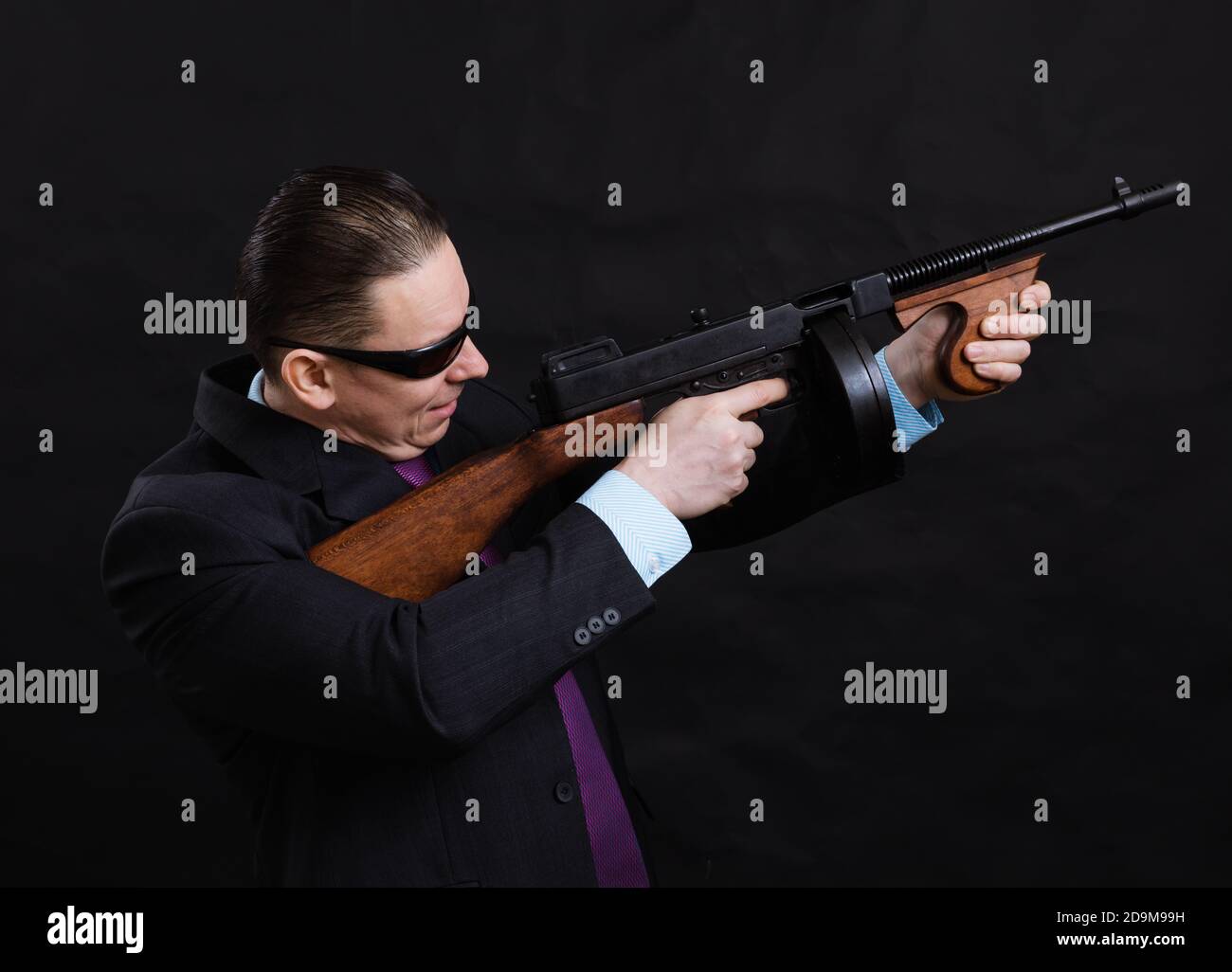 Gangster mature en lunettes de soleil vêtu en costume avec un fusil tommy sur fond noir Banque D'Images