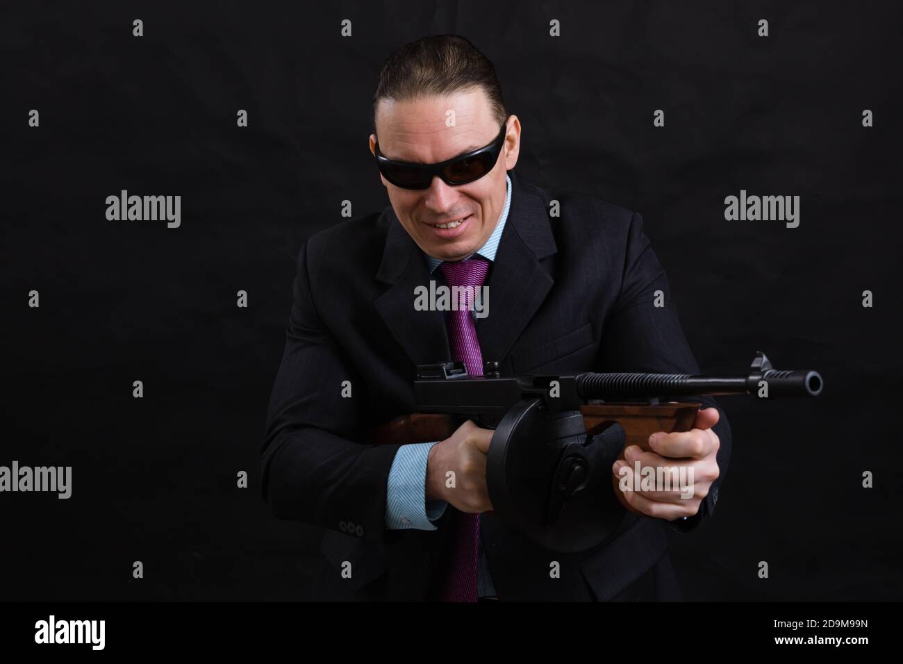 Gangster mature en lunettes de soleil vêtu en costume avec un fusil tommy sur fond noir Banque D'Images