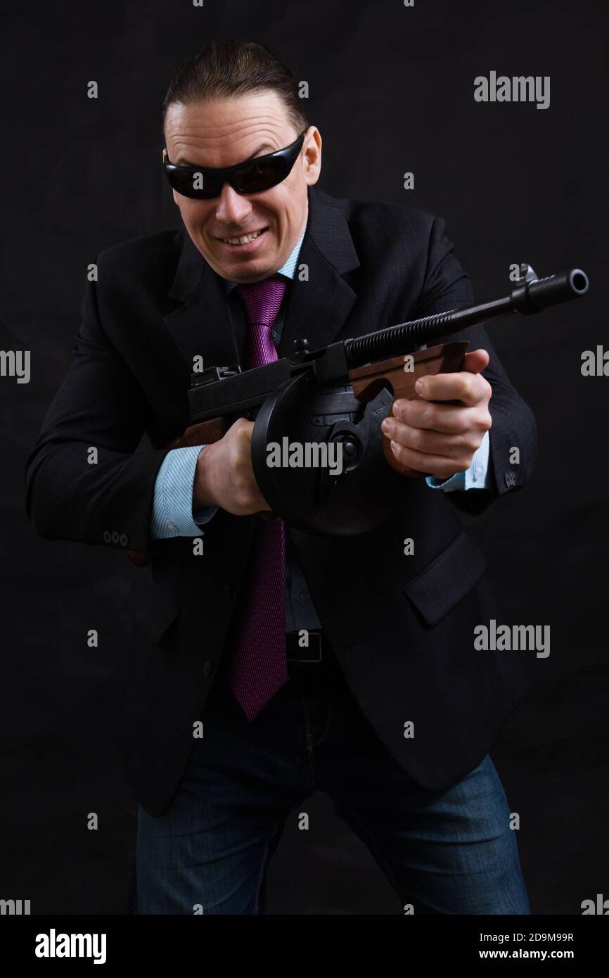 Gangster mature en lunettes de soleil vêtu en costume avec un fusil tommy sur fond noir Banque D'Images