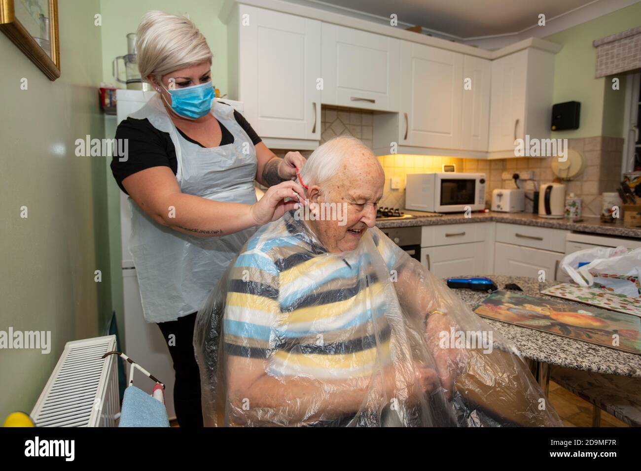 Homme âgé dans les années quatre-vingt ayant ses cheveux coupés par un coiffeur portant un EPI pour la protection contre le coronavirus tout en travaillant à distance, Angleterre, Royaume-Uni Banque D'Images