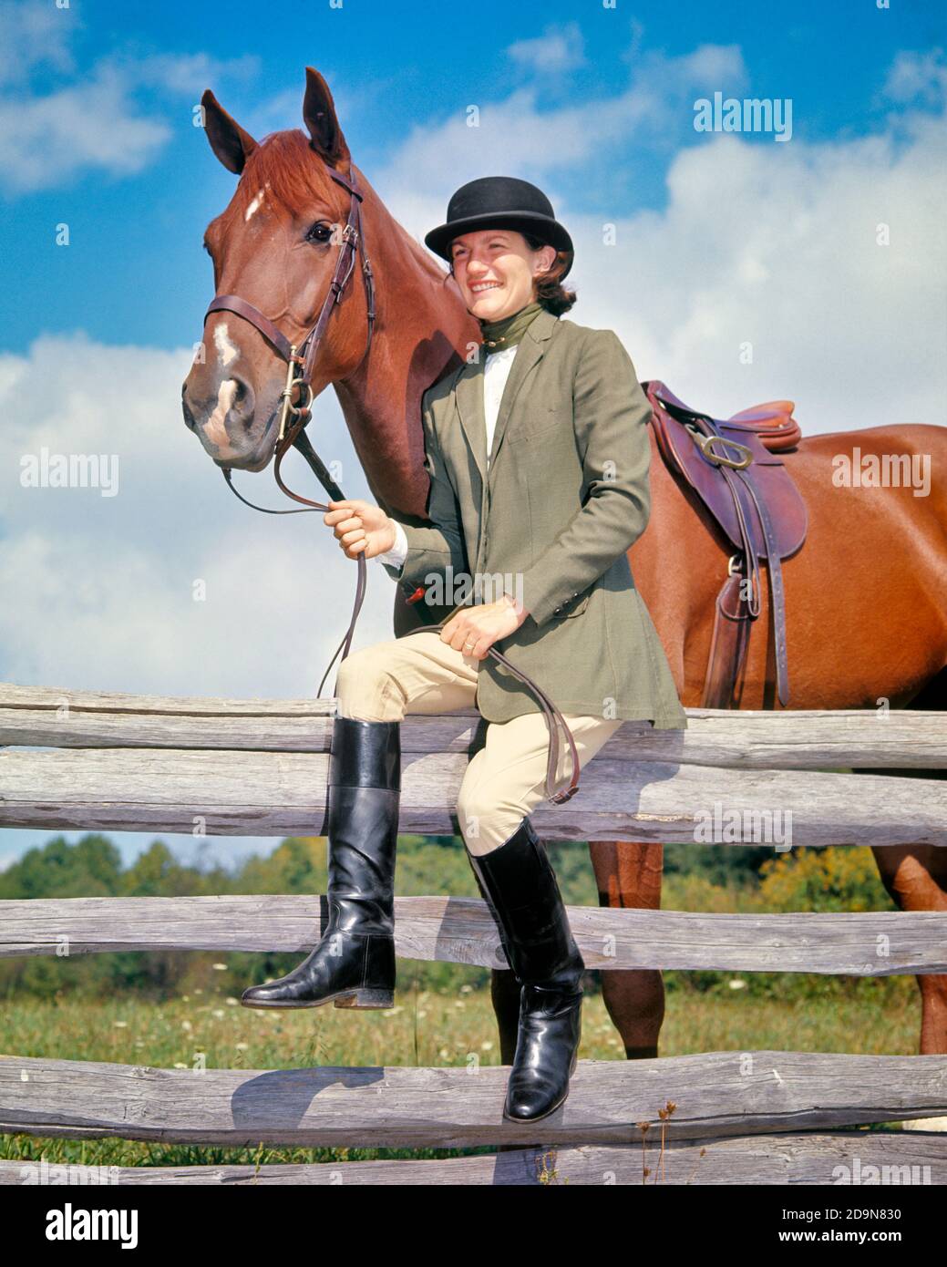 FEMME DES ANNÉES 1970 PORTANT LE STYLE DE BOWLER CHAPEAU VESTE D'ÉQUITATION  CULOTTES BOTTES ASSIS SUR LA CLÔTURE DE RAIL TENANT LE CHEVAL DE REINS AVEC  LA SELLE ANGLAISE - KH3500 HAR001