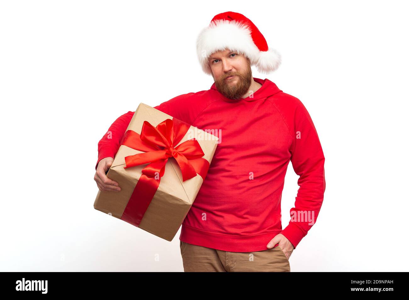 Un homme brutal avec une barbe rouge dans un chapeau de père noël regarde la caméra et tient une boîte cadeau avec un ruban rouge dans ses mains. Studio shot Isolate, chris Banque D'Images