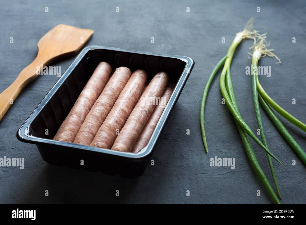 Dans un récipient en plastique sur fond gris, plusieurs saucisses sous forme de produits semi-finis sont disponibles. Près de se trouve un frais les plumes d'oignons verts Banque D'Images