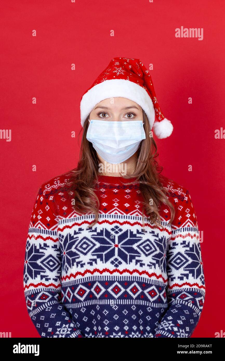 Jeune femme en masque médical, chapeau du Père Noël et chandail à motifs isolés sur fond rouge Banque D'Images