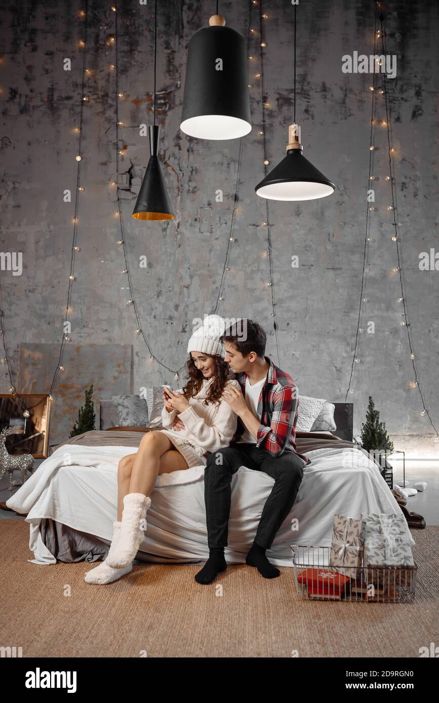 Photo pleine longueur de deux personnes gaies utiliser un homme de téléphone portable Afficher les réductions saisonnières de Noël Félicitations avec Noël assis sur le lit intérieur avec une atmosphère de décoration de nouvel an Banque D'Images