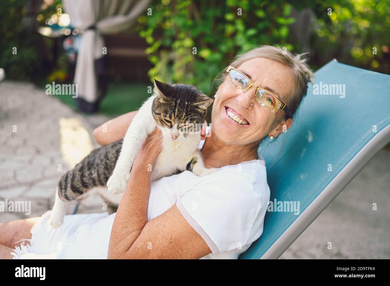 Heureuse femme âgée souriante dans des verres se détendre dans le jardin d'été à l'extérieur embrassant domestique tabby chat. Concept de vieux retraités et animaux de compagnie Banque D'Images