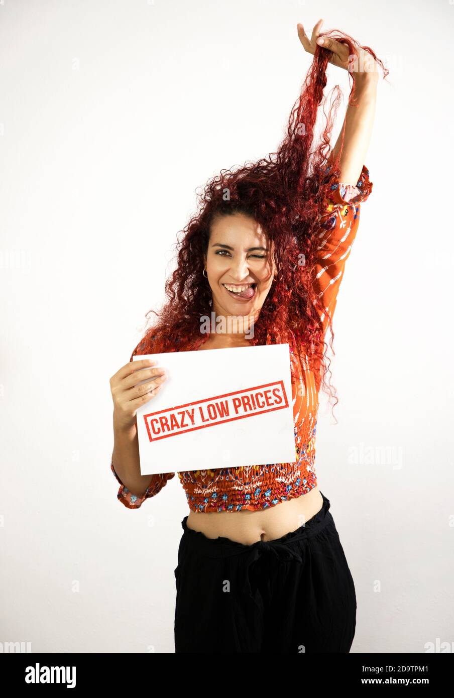 Très heureuse fille souriante avec la langue dehors soulevant ses cheveux avec une main et tenant un signe fou de bas prix avec l'autre main. Jeune femme avec Banque D'Images