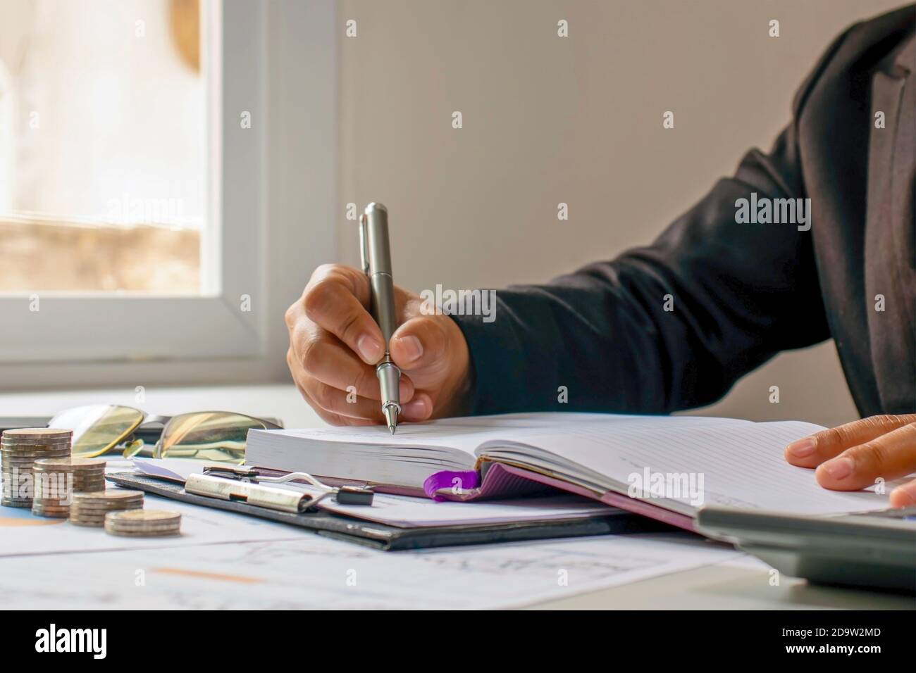 Une femme écrit dans un bloc-notes sur le bureau avec une pile de pièces à côté de la fenêtre de la maison et des idées éducatives. Banque D'Images