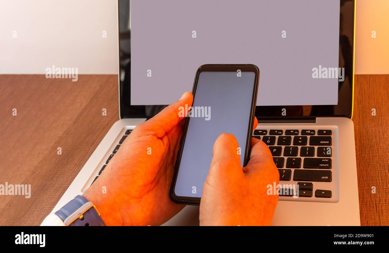 Mains d'un homme qui manipule un téléphone cellulaire attaché à un ordinateur portable avec un écran vierge sur un bureau en bois avec un éclairage chaud, maquette de téléphone et d'ordinateur portable Banque D'Images