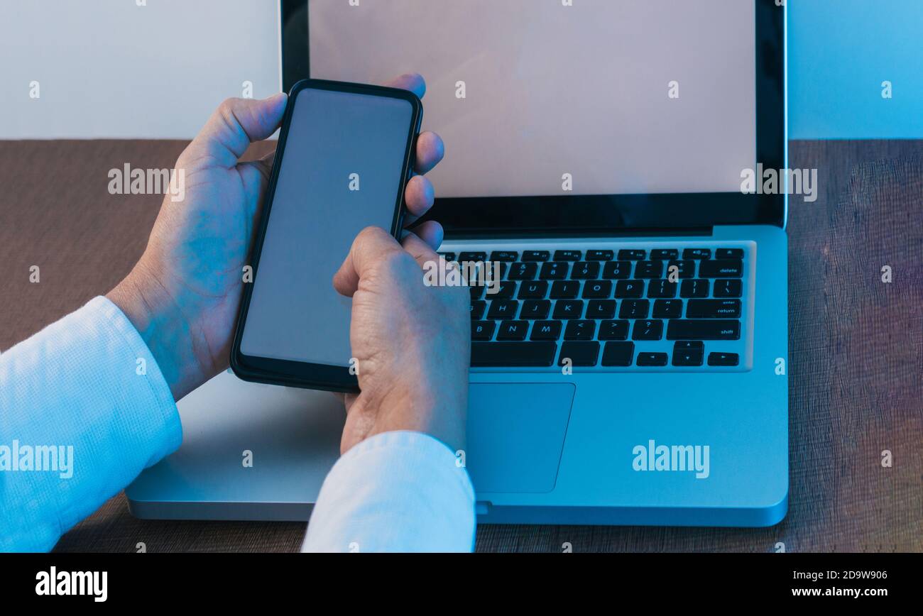 mains d'un homme qui manipule un téléphone cellulaire attaché à un ordinateur portable avec un écran blanc sur un bureau en bois avec un éclairage bleu, maquette d'ordinateur portable et smartphone Banque D'Images