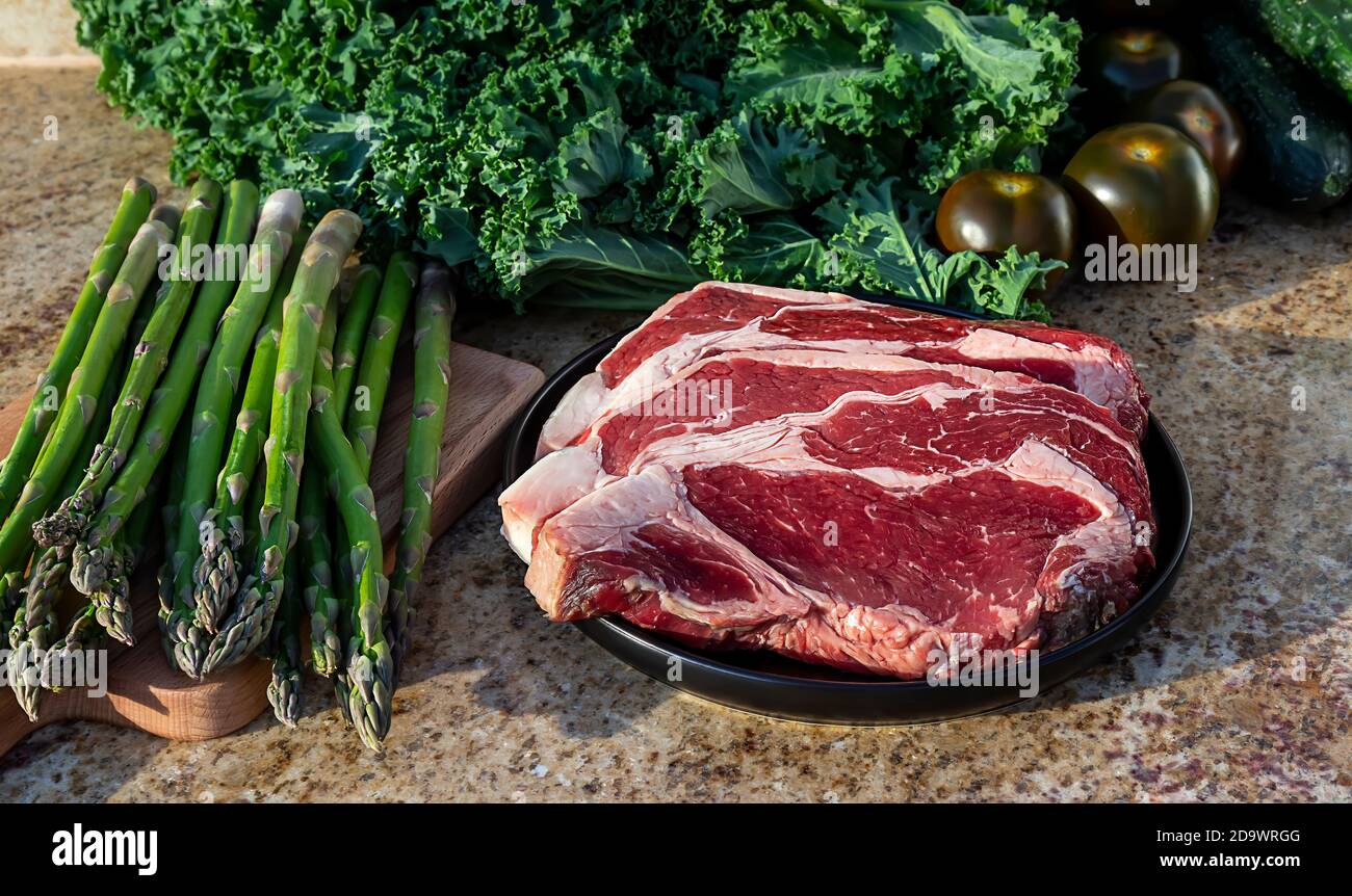 Viande crue, steak de bœuf sur une assiette noire avec chou vert, asperges, broccolini et tomates en arrière-plan. Banque D'Images