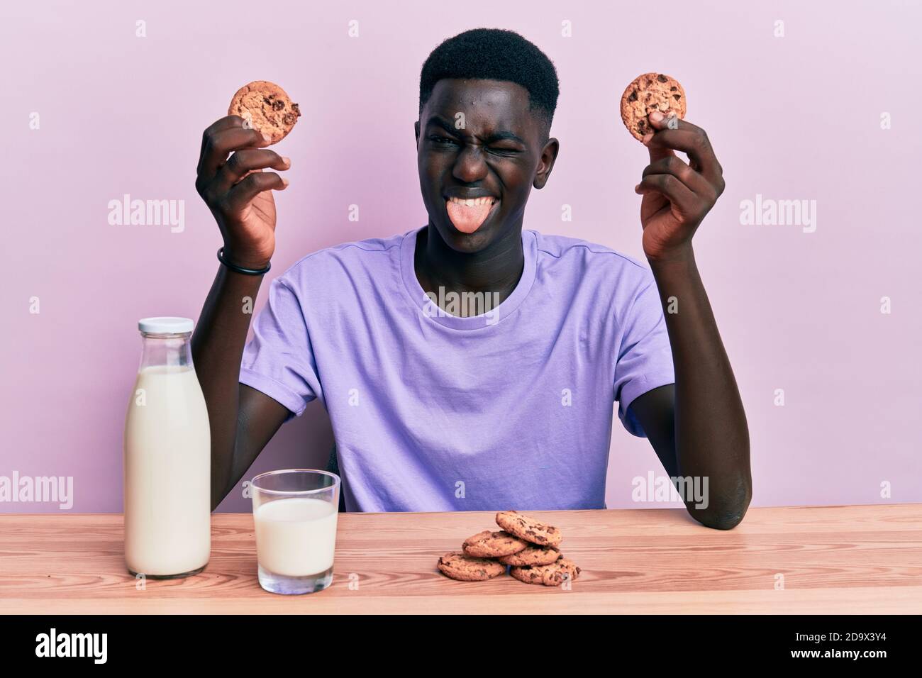 Un jeune homme afro-américain boit un verre de lait frais avec des biscuits qui collent la langue heureux avec l'expression drôle. Banque D'Images