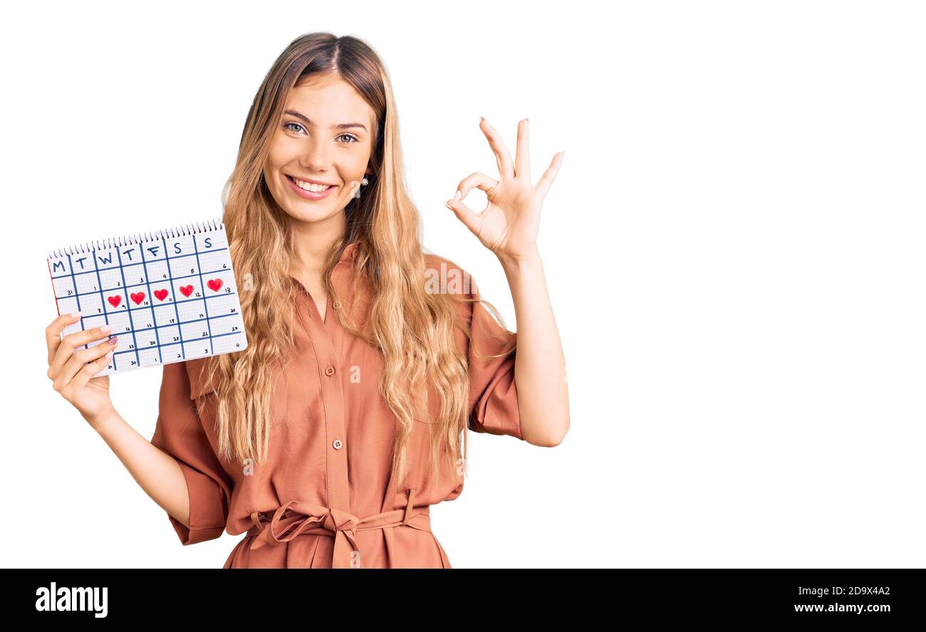 Belle femme caucasienne avec cheveux blonds tenant le calendrier cardiaque faisant signe ok avec les doigts, souriant sympathique gestante excellent symbole Banque D'Images