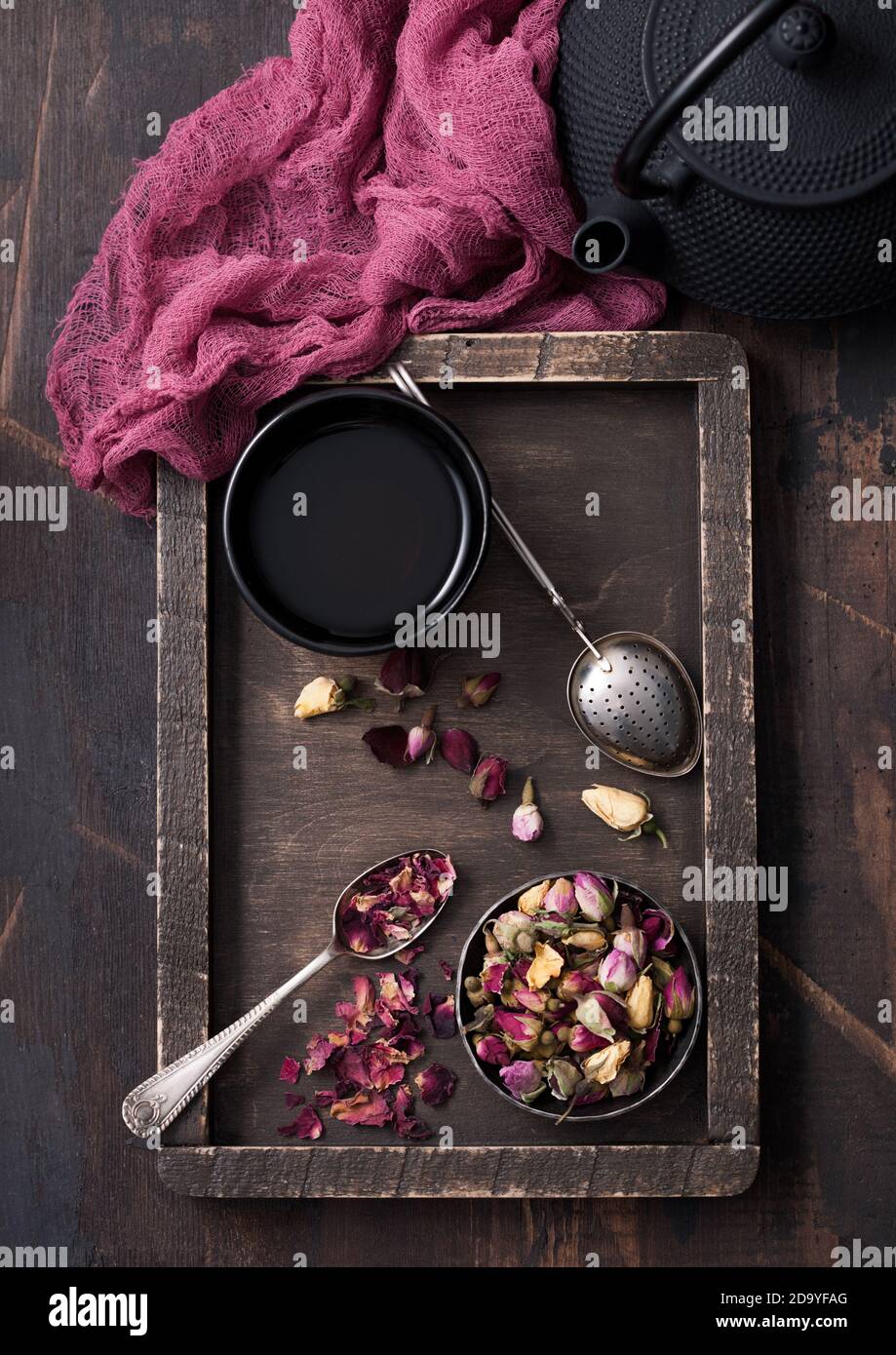 Rose bourgeons thé dans une boîte en bois avec une cuillère et un infuseur de passoire et des tasses en fer avec théière et tissu rouge sur fond de bois. Vue de dessus Banque D'Images