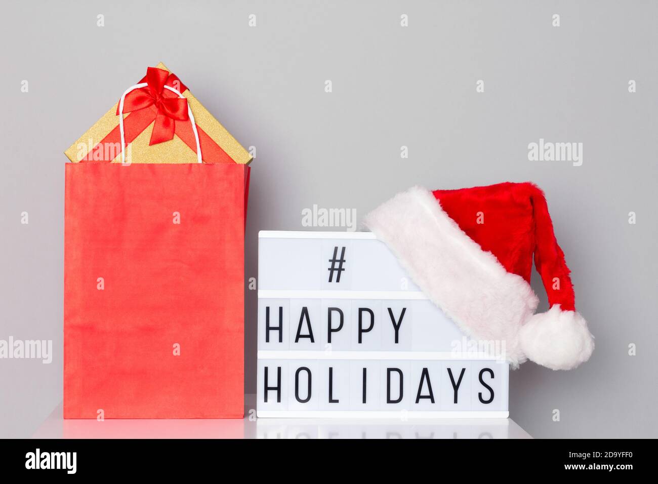 Visionneuse avec hashtag hashtag happy Holidays avec chapeau de père Noël et Sac à provisions en papier rempli de cadeaux de Noël sur table blanche Banque D'Images
