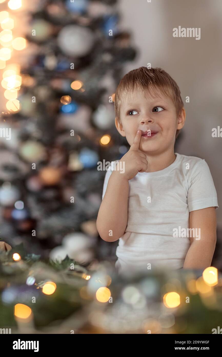 Un beau garçon de 4-5 ans est assis ou debout près de l'arbre de Noël. Il rêve de vacances et de cadeaux, fait un vœu. Le concept du wi Banque D'Images