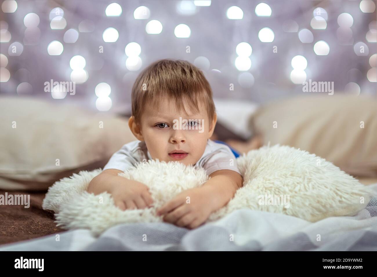 Un beau garçon de 4-5 ans est allongé sur un lit sur un fond de guirlandes étincelantes. Ambiance chaleureuse des vacances de Noël. Le concept Banque D'Images