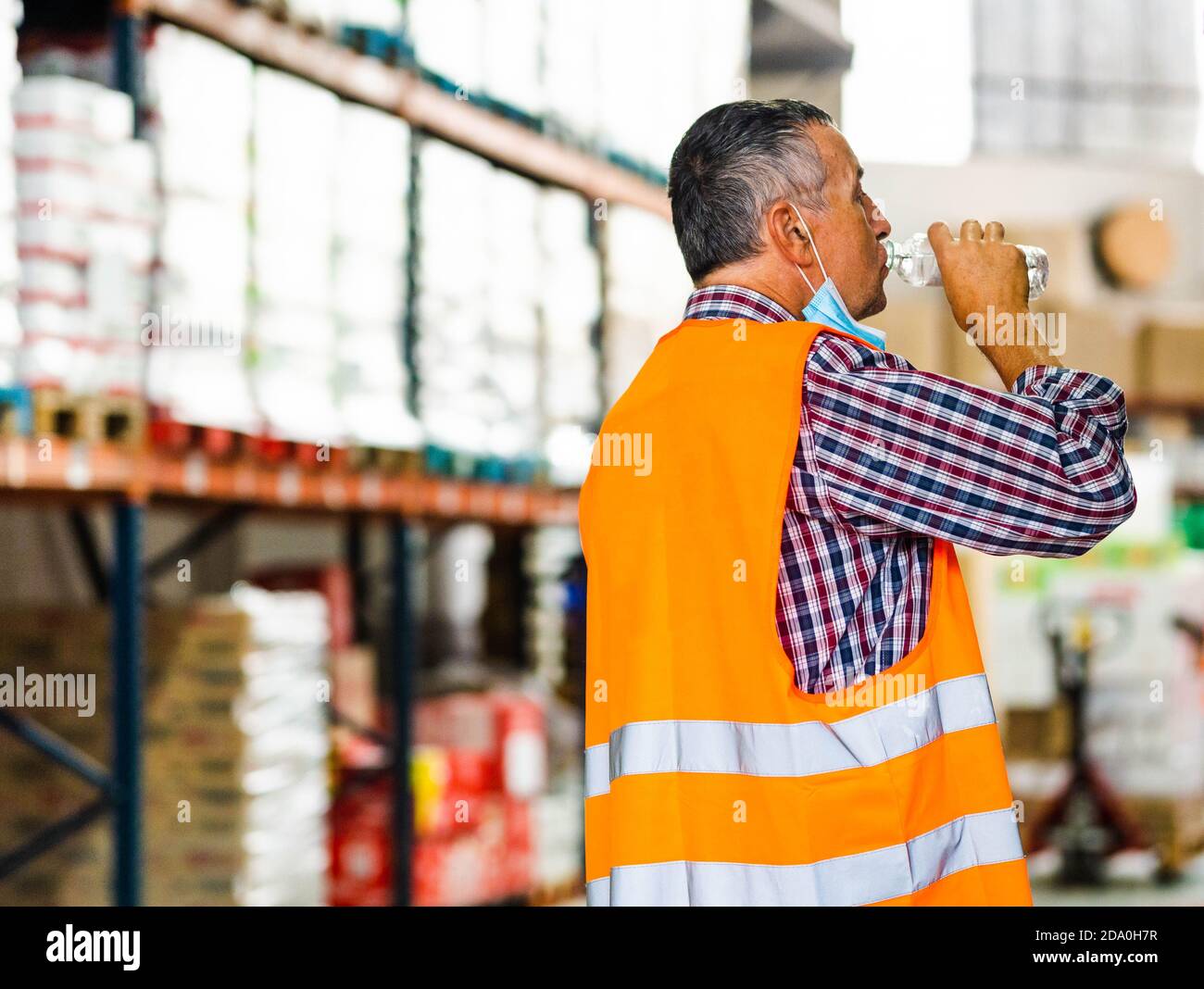 Vue latérale adulte homme travailleur dans un gilet orange haute visibilité et masque abaissé eau potable de bouteille en plastique en position debout près des étagères dans l'entrepôt Banque D'Images