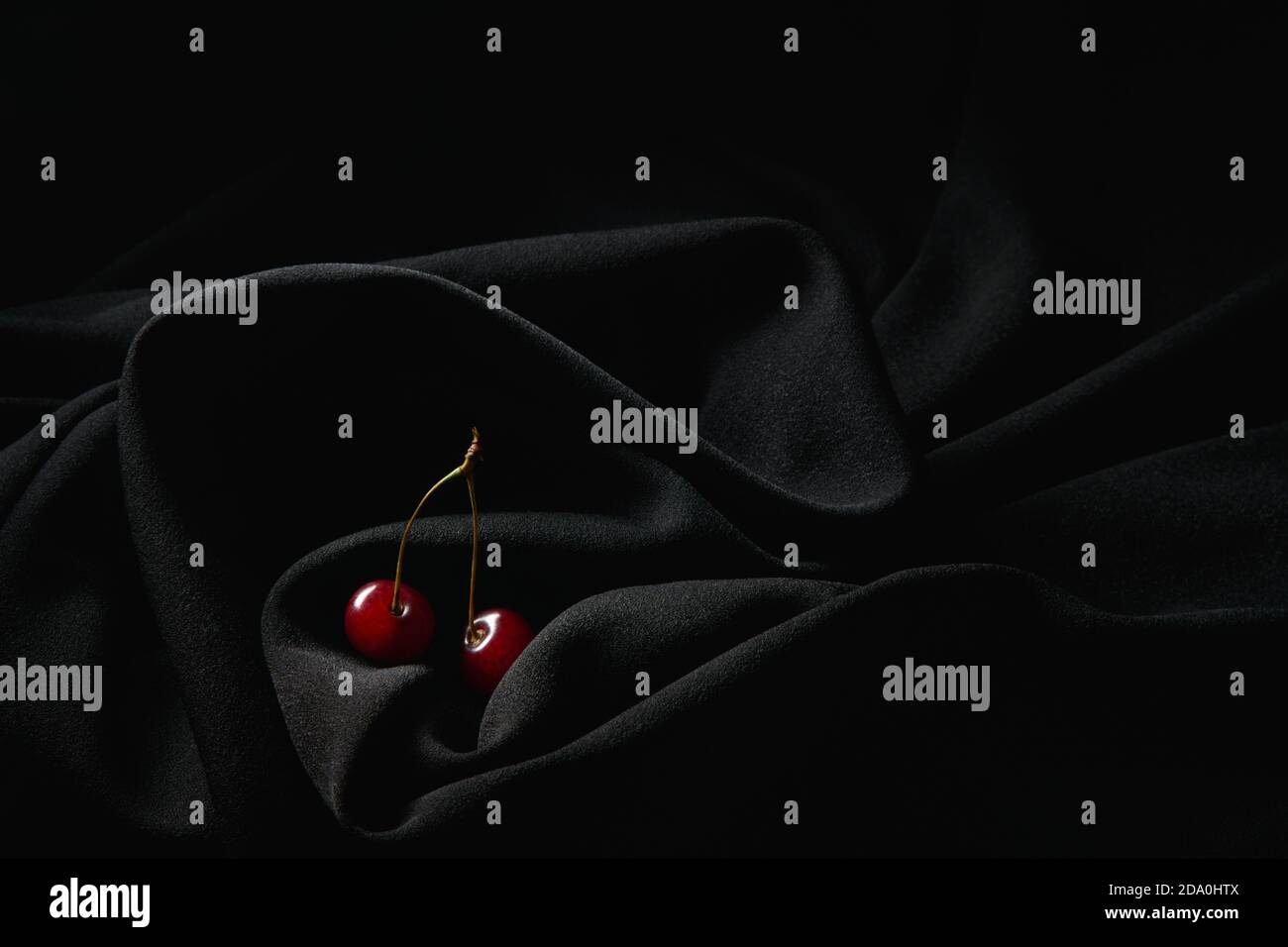 Petites cerises rouges sur les tiges minces placées sur le noir foncé chiffon froissé en studio Banque D'Images