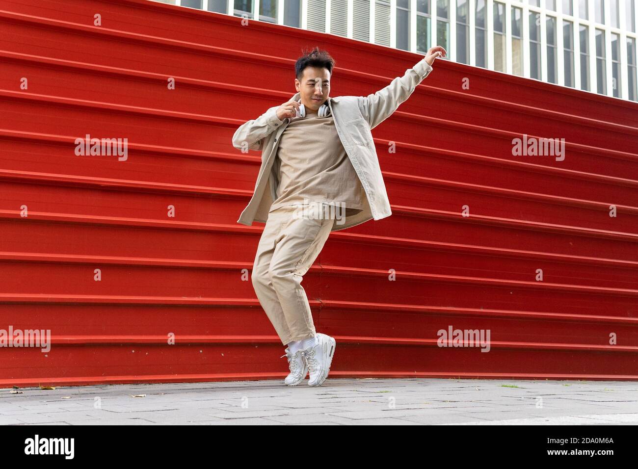 Homme asiatique plein de corps joyeux en tenue décontractée à l'écoute chanson préférée dans un casque sans fil tout en dansant le long du métal rouge clôture Banque D'Images