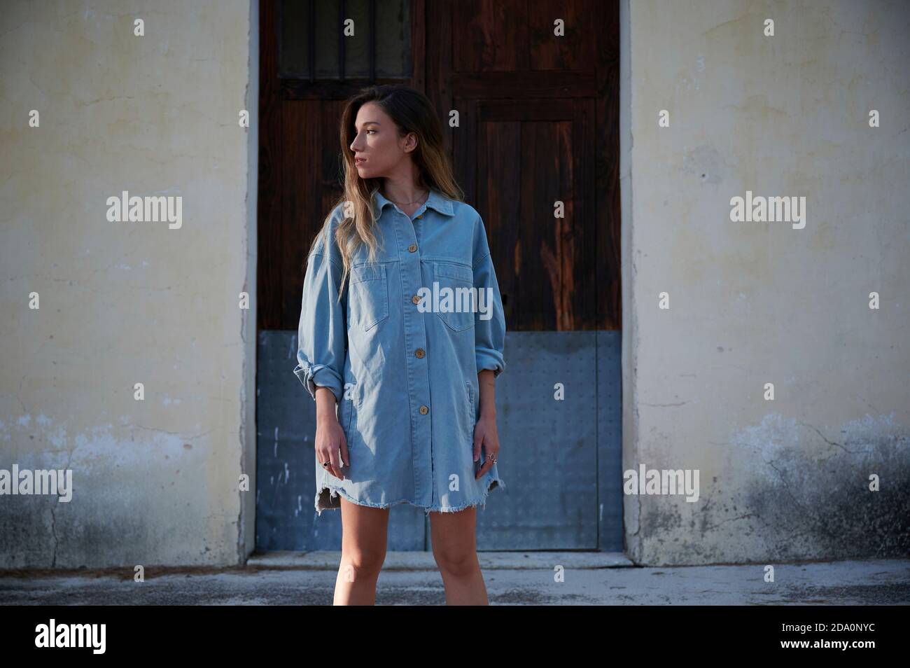 Jeune femme cintrée avec robe en denim élégante et bottes debout sur pelouse herbacée couverte de givre contre la maison rurale abîmé en automne Banque D'Images