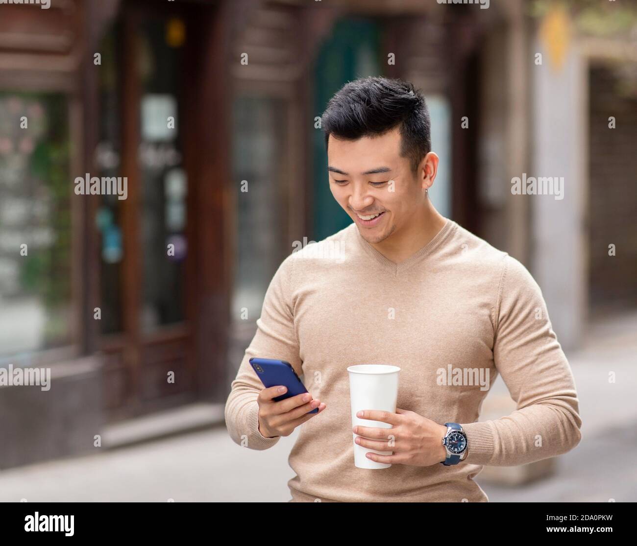 Homme asiatique souriant debout dans la rue avec une boisson chaude aromatique pour lire des messages sur les réseaux sociaux via un téléphone mobile téléphone Banque D'Images