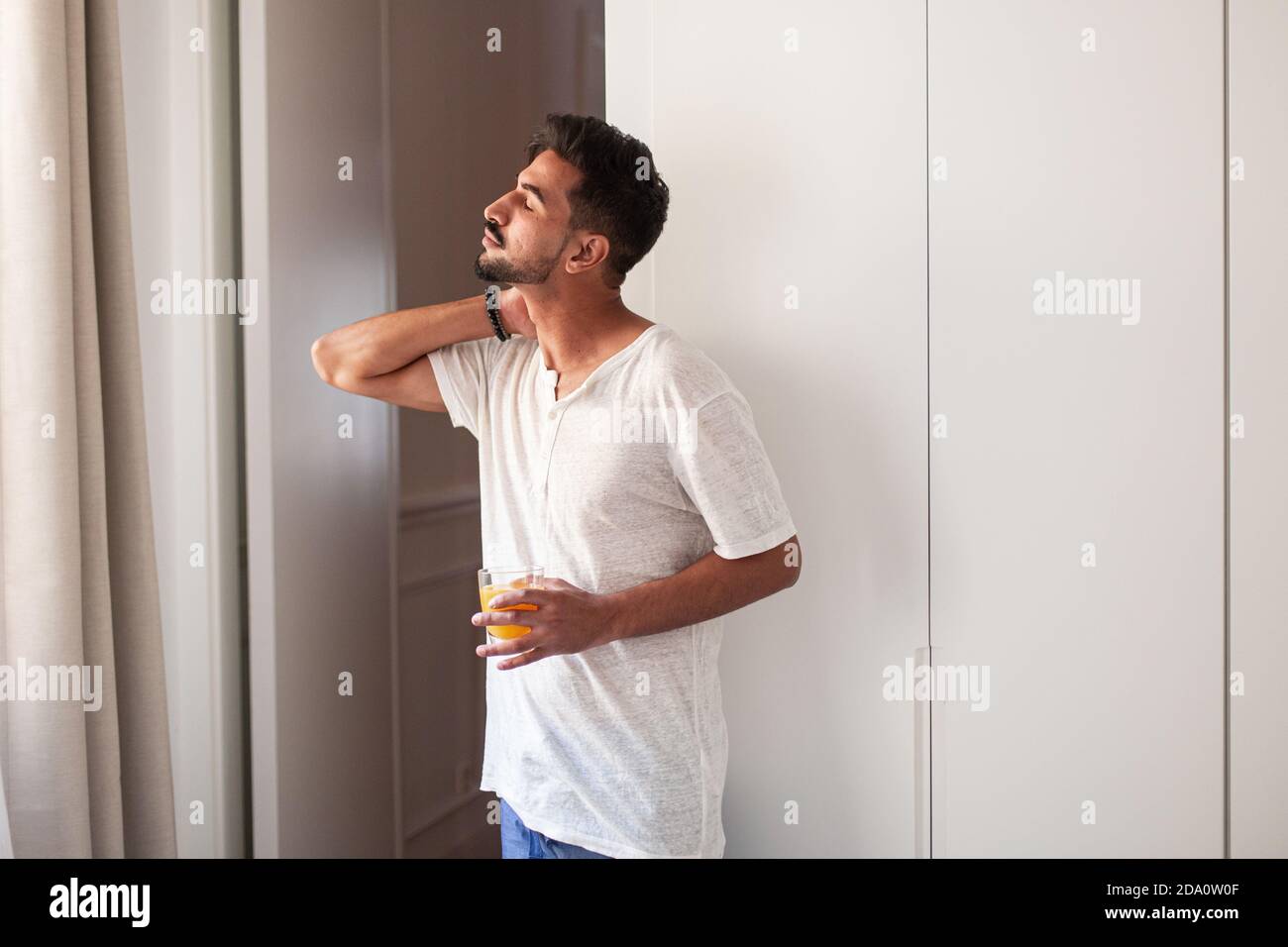 Vue latérale d'un jeune homme dans des vêtements décontractés qui boivent frais pressez et observez la rue tout en se tenant près de la fenêtre dans moderne appartement le matin avec oeil Banque D'Images