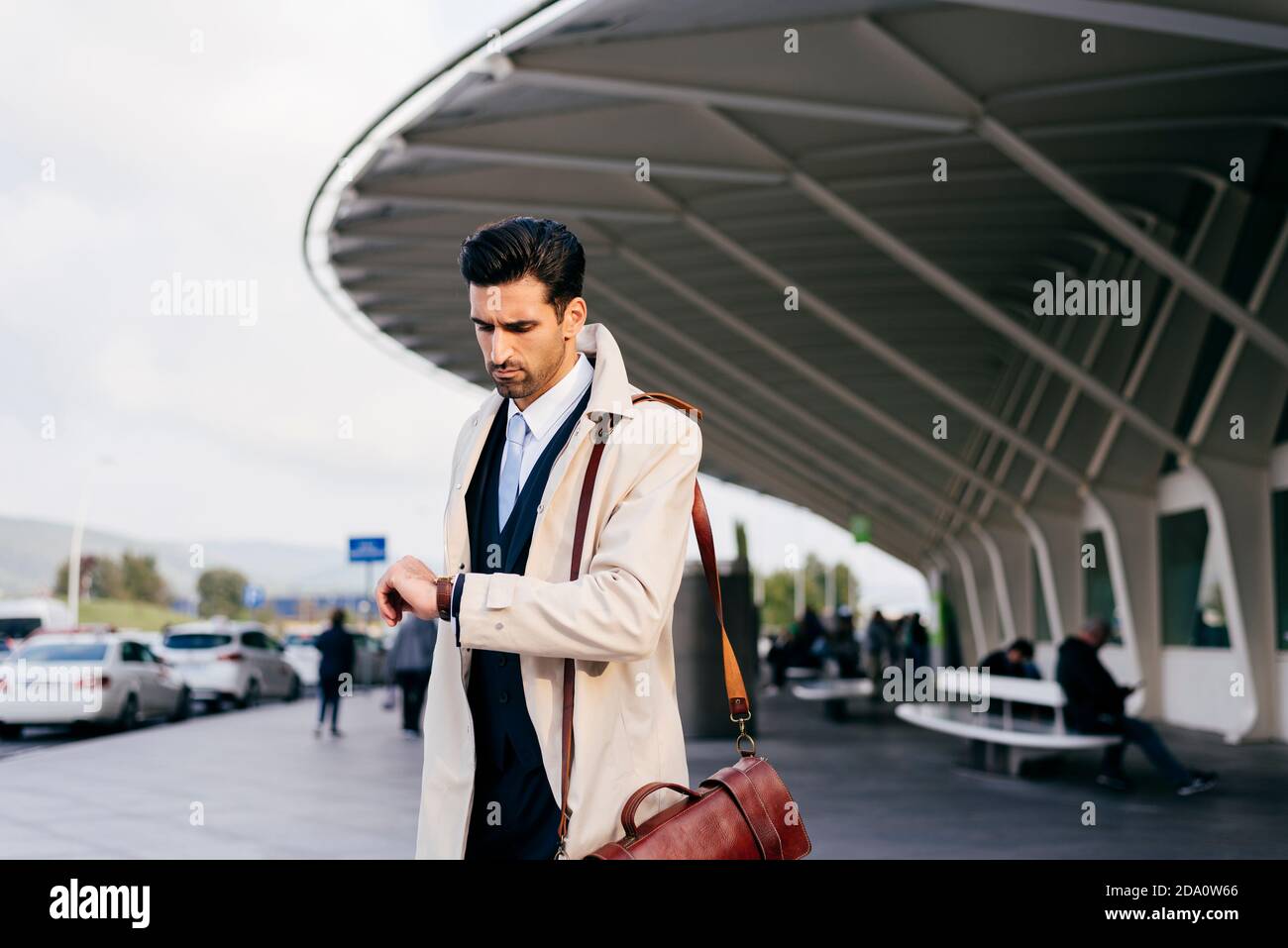 homme aux cheveux foncés en costume et manteau regardant votre montre tout en se tenant près du terminal de l'aéroport le jour Banque D'Images