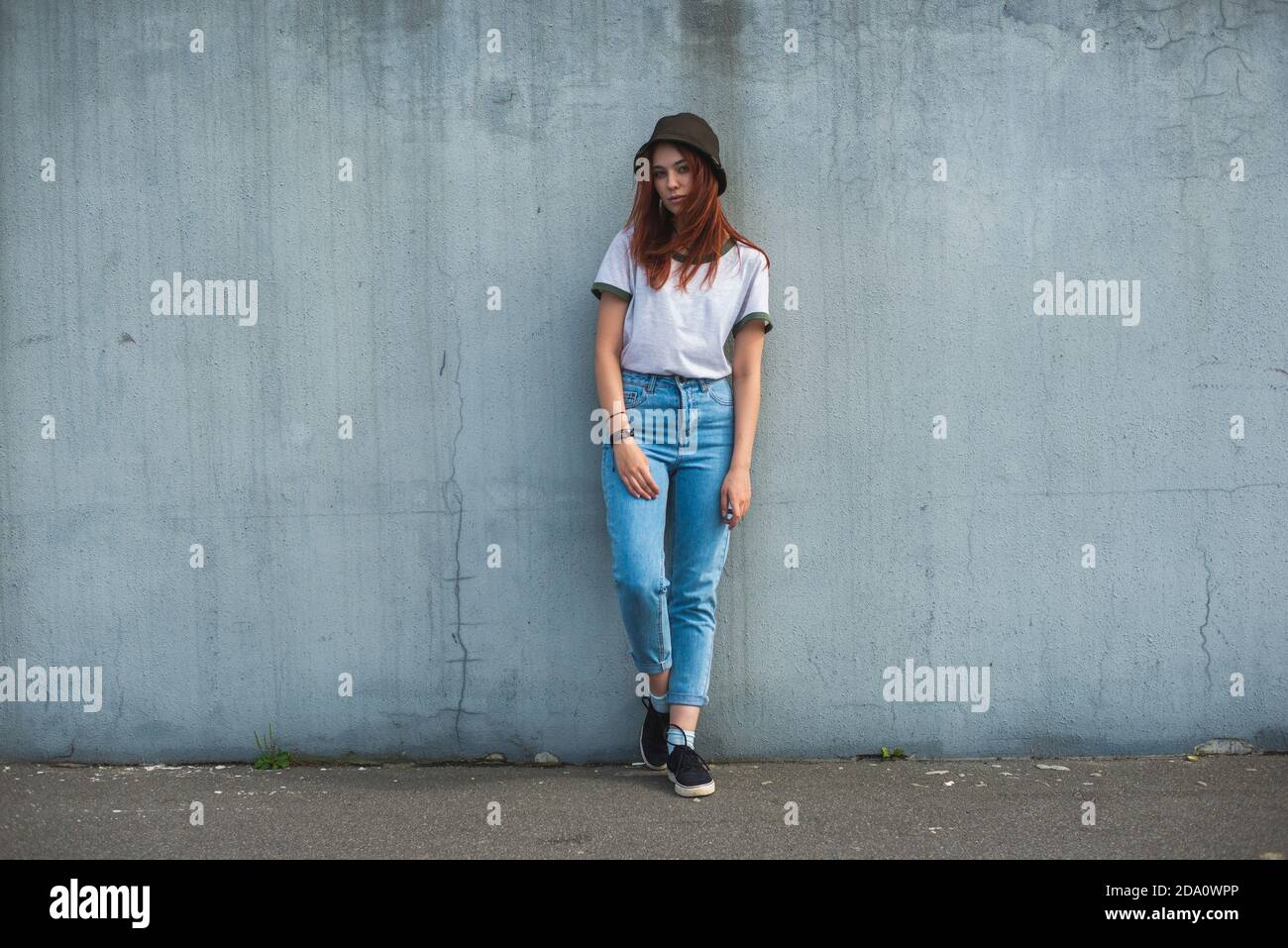 Tout le corps de l'adolescence tendance rouge cheveux femelle élève dans tenue décontractée et chapeau incliné vers l'arrière sur béton gris mur sur la rue urbaine Banque D'Images